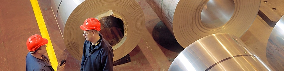 two factory workers talking next to large rolls of steel